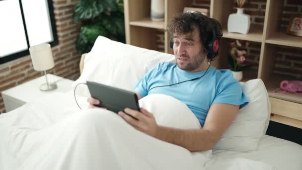 Young Hispanic Man Using Touchpad Headphones Lying Bed Bedroom — Stockvideo