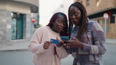 Two african american women using smartphone and credit card at street