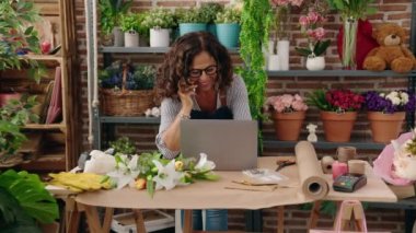 Middle age woman florist talking on smartphone using laptop at flower shop