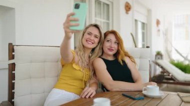 Evdeki terasta oturan iki kadın akıllı telefondan selfie çekiyor.
