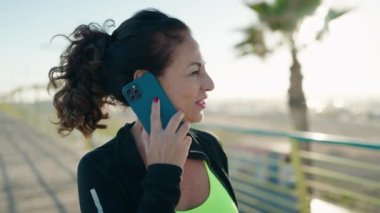 Orta yaşlı kadın spor kıyafeti giyiyor ve caddede akıllı telefondan konuşuyor.
