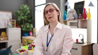 Young blonde woman preschool teacher sitting on chair speaking at kindergarten