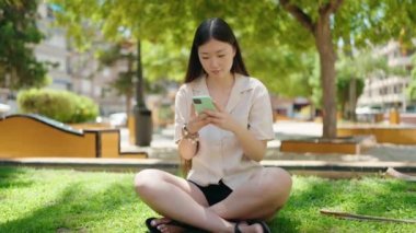 Parkta çimlerin üzerinde oturan akıllı telefon kullanan Çinli bir kadın.