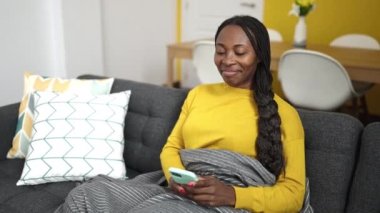 African woman using smartphone sitting on sofa at home