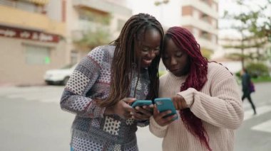 Two african american women smiling confident using smartphone at street