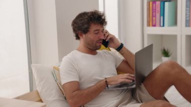 Young hispanic man using laptop talking on smartphone at bedroom