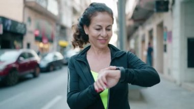 Orta yaşlı kadın spor kıyafeti giyiyor. Gülümsüyor. Kendine güvenen kronometreye bakıyor.