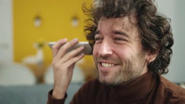 Genç İspanyol adam evdeki koltukta oturmuş akıllı telefondan sesli mesaj dinliyor.