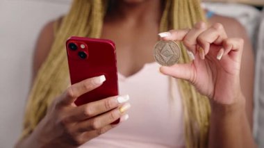 African american woman holding ethereum classic crypto currency using smartphone at home