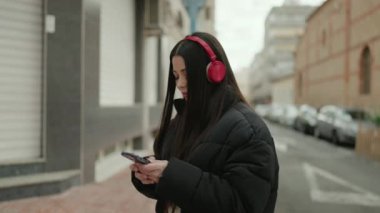 Young hispanic woman listening to music using smartphone at street