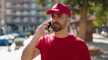 Genç İspanyol teslimatçı çantayla parkta akıllı telefondan konuşuyor.