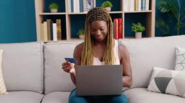 Evdeki koltukta oturan Afrikalı Amerikalı kadın dizüstü bilgisayar ve kredi kartı kullanıyor.