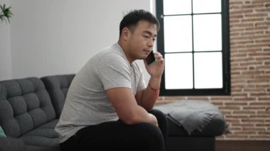 Genç Çinli adam evde endişeli bir ifadeyle akıllı telefondan konuşuyor.