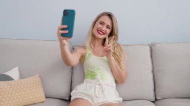 Young beautiful hispanic woman make selfie by smartphone sitting on sofa at home