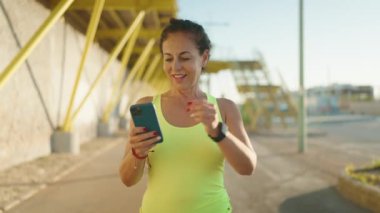 Middle age woman wearing sportswear using smartphone looking stopwatch at street