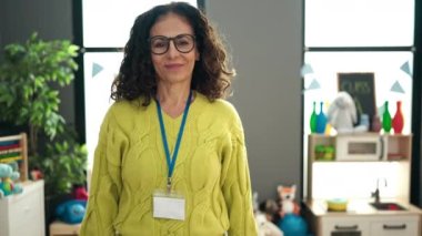 Orta yaşlı İspanyol bir anaokulu öğretmeni. Anaokulunda kollarını kavuşturup gülümsüyor.