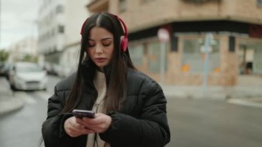 Young hispanic woman listening to music using smartphone at street