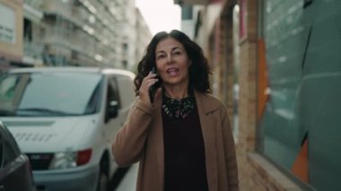 Orta yaşlı İspanyol kadın gülümsüyor. Akıllı telefondan konuşuyor. Sokakta yürüyor.