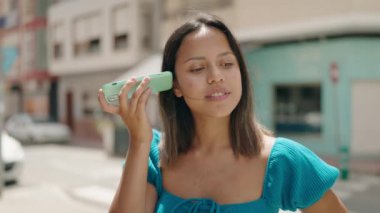 Young hispanic woman smiling confident listening audio message by the smartphone at street