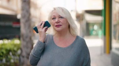 Middle age blonde woman listening audio message by the smartphone at street