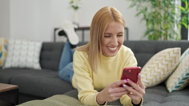 年轻的金发女人用智能手机躺在沙发上 — 图库视频影像