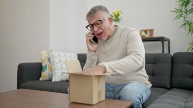 Orta yaşlı, kır saçlı, akıllı telefondan konuşan evde paket bekleyen bir adam.