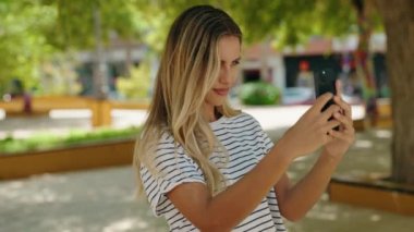 Kendine güvenen genç sarışın kadın parkta akıllı telefondan fotoğraf çekiyor.