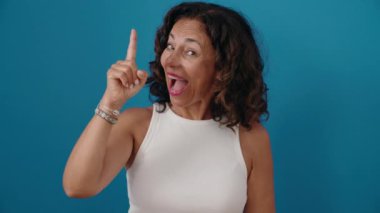 Orta yaşlı kadın kendinden emin gülümsüyor. Mavi arka planda parmaklarıyla fikir jesti yapıyor.