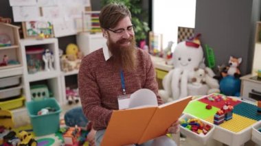 Young redhead man preschool teacher reading book sitting on chair at kindergarten
