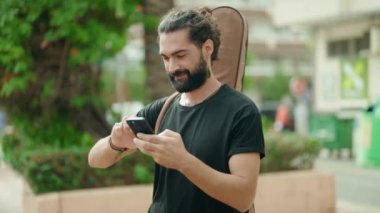 Genç İspanyol müzisyen parkta gitar çantası ile akıllı telefon kullanıyor.