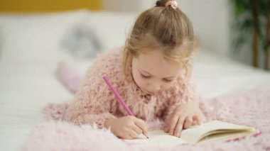 Adorable blonde girl drawing on notebook lying on bed at bedroom