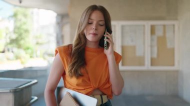 Üniversitede elinde laptop ve kitapla akıllı telefondan konuşan genç bir kız öğrenci.