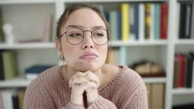 Young beautiful hispanic woman student sitting on table with doubt expression at library university