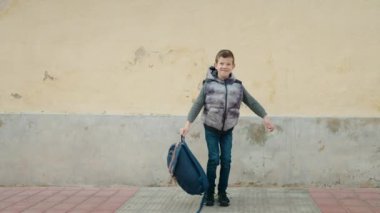 Sırt çantası taşıyan sarışın bir çocuk sokakta zıplıyor.