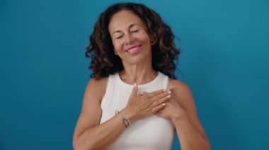 Orta yaşlı kadın gülümsüyor. Elleri kalbimde, mavi arka planda duruyor.