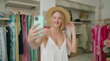 Genç sarışın kadın müşteri elbise mağazasında akıllı telefondan selfie çekiyor.