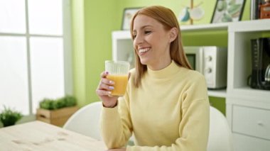 Young blonde woman drinking orange juice sitting on table at home