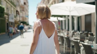 Genç sarışın kadın gülümsüyor. Kollarını kavuşturup caddede duruyor.