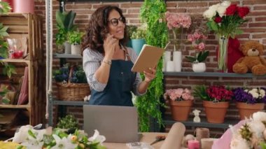 Middle age woman florist talking on smartphone reading book at flower shop