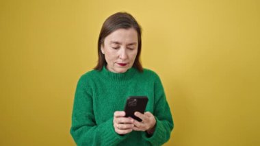 Gri saçlı, akıllı telefon kullanan, izole edilmiş sarı arka planda gülümseyen olgun İspanyol kadın.