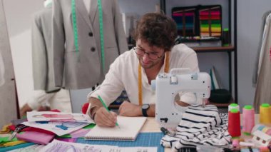 Genç İspanyol terzi gülümseyerek kıyafet fabrikasında deftere çizim yapıyor.