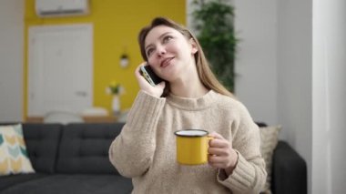 Young blonde woman talking on smartphone drinking coffee at home