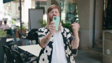 Young redhead man using smartphone with winner expression at coffee shop terrace