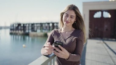 Genç sarışın kadın, kendinden emin gülümsüyor. Deniz kenarındaki akıllı telefonun yanında özçekim yapıyor.