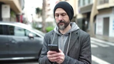 Genç kel adam akıllı telefon kullanıyor ve sokakta ciddi bir yüz ifadesi var.