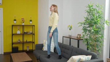 Genç sarışın kadın gülümsüyor evdeki kanepede dans ediyor.