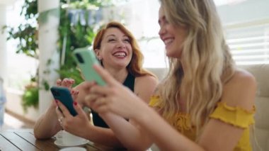 Two women using smartphones and drinking coffee sitting on table at home terrace