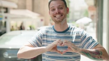 Young hispanic man smiling confident doing heart gesture with hands at street