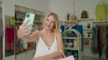 Alışveriş torbaları tutan genç sarışın kadın giyim mağazasında akıllı telefondan selfie çekiyor.