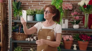 Orta yaşlı kadın çiçekçi çiçekçide akıllı telefondan selfie çekiyor.